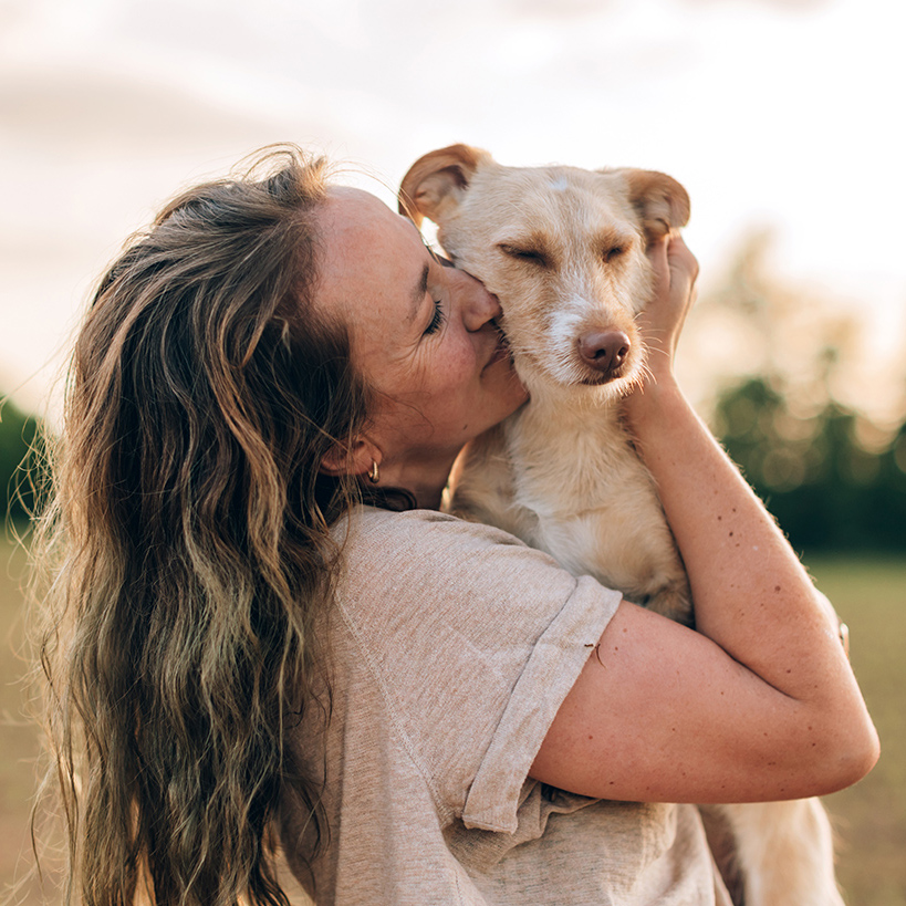 woman with long hair hugs and kisses a small, tan dog in a sunlit field